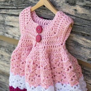 May include: A pink crocheted baby dress with white and pink trim. The dress has two red buttons on the front.