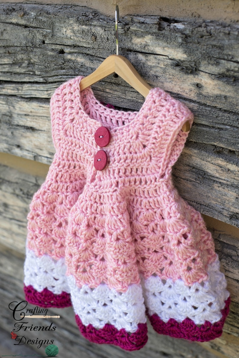 May include: A pink crocheted baby dress with white and pink trim. The dress has two red buttons on the front.