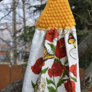 May include: A hanging kitchen towel with a yellow crocheted top and a button fastening. The white towel displays red poppies, butterflies, and the text "Poppies and Butterflies".