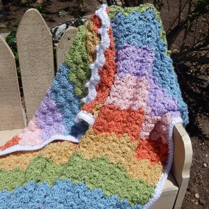 May include: A multi-colored crocheted baby blanket with a white crocheted border. The blanket is made in a variety of pastel colors, including pink, blue, green, and yellow.