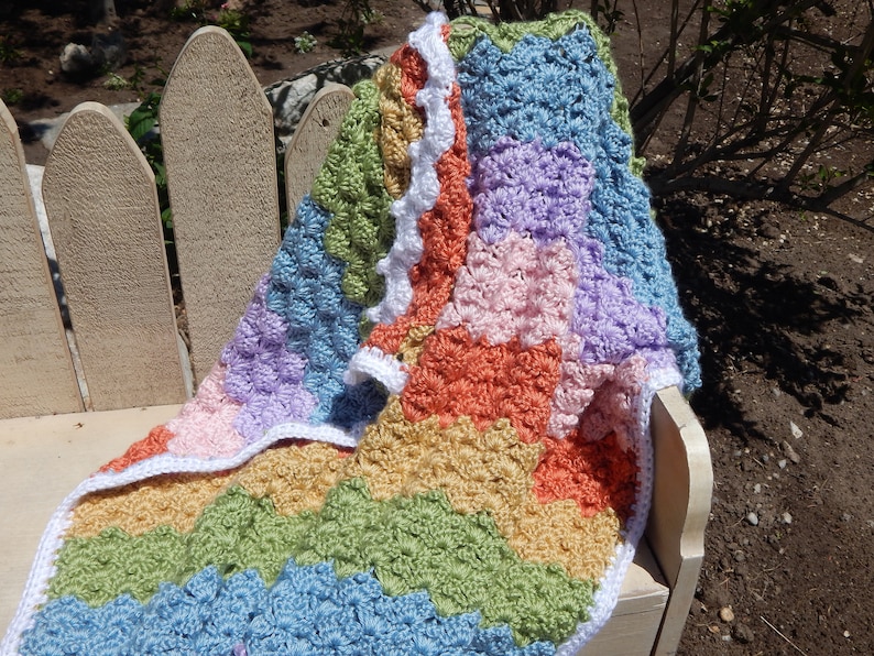 May include: A multi-colored crocheted baby blanket with a white crocheted border. The blanket is made in a variety of pastel colors, including pink, blue, green, and yellow.
