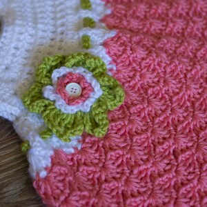 Peut inclure: Une robe de b&eacute;b&eacute; en crochet avec un corsage blanc et une jupe rose. La robe est orn&eacute;e d'une applique de fleur verte et blanche.