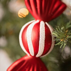 May include: Red and white candy cane Christmas ornament hanging on a green Christmas tree. The ornament is shaped like a candy cane with a red and white striped center and red ends.
