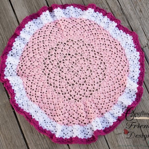 May include: A pink and white crocheted baby blanket with a pink and white crocheted border. The blanket is round and has a scalloped edge. The blanket is on a wooden surface.