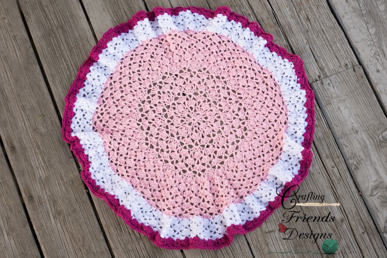 May include: A pink and white crocheted baby blanket with a pink and white crocheted border. The blanket is round and has a scalloped edge. The blanket is on a wooden surface.