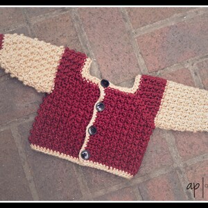 May include: A crocheted baby cardigan with a burgundy body and cream coloured sleeves and trim. The cardigan has a button closure.