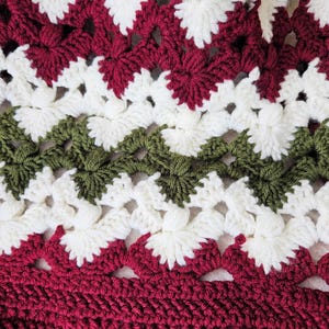 May include: A crocheted blanket with a repeating pattern of white, red, and green flowers. The blanket is made with a soft yarn and has a textured surface.