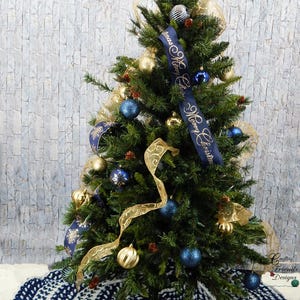 May include: A blue and white crocheted Christmas tree skirt with a white background and blue circles. A small Christmas tree decorated with gold and blue ornaments and a ribbon that says "Merry Christmas" is in the background.