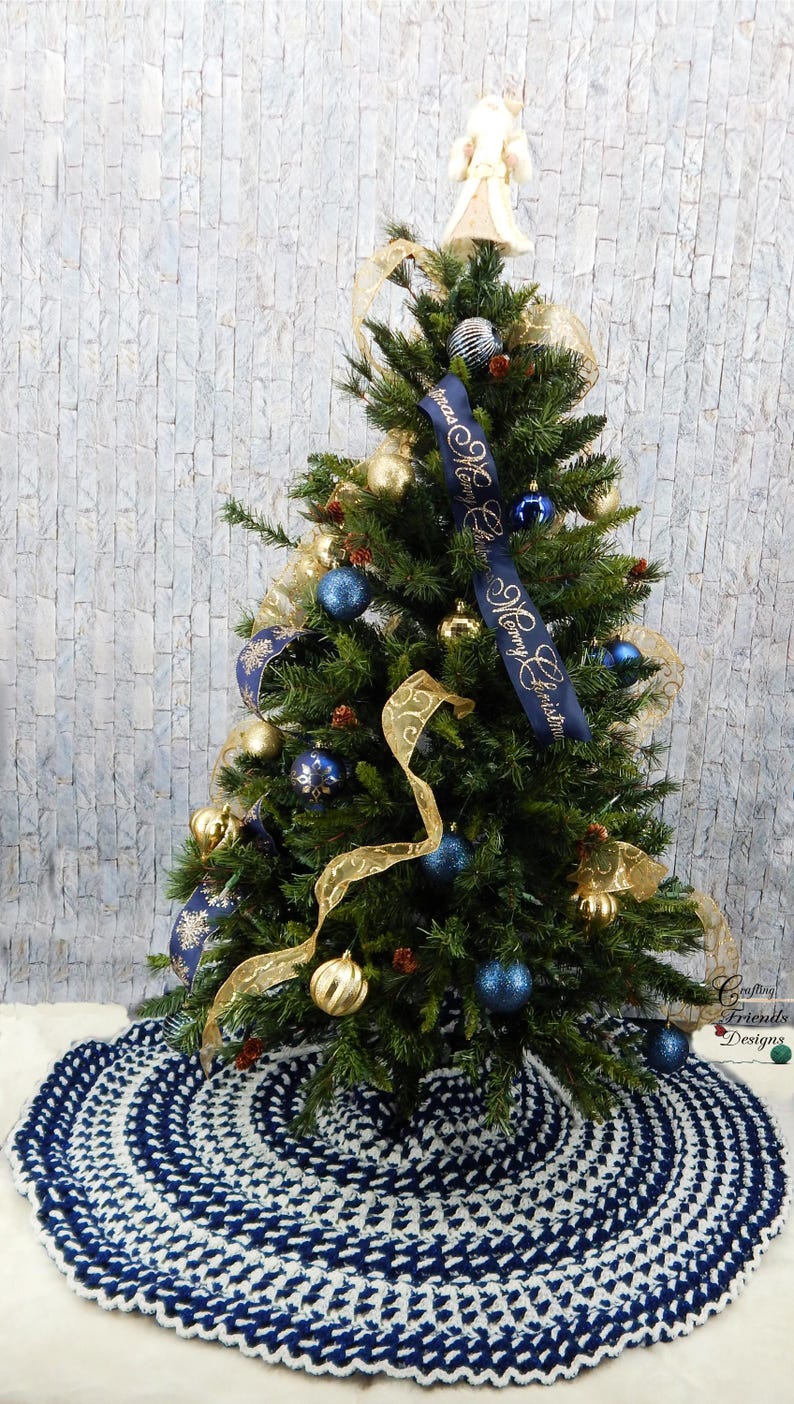 May include: A blue and white crocheted Christmas tree skirt with a white background and blue circles. A small Christmas tree decorated with gold and blue ornaments and a ribbon that says "Merry Christmas" is in the background.