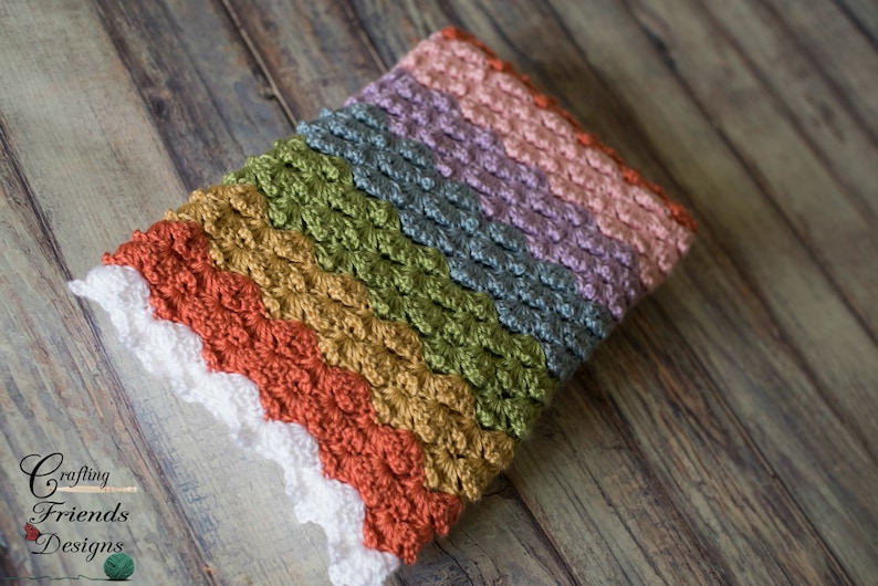 May include: A multi-colored crocheted baby blanket with a white crocheted border. The blanket is folded in half and lying on a wooden surface.