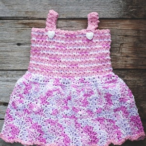 May include: A pink and white crocheted baby dress with a scalloped hem and two white heart-shaped buttons.
