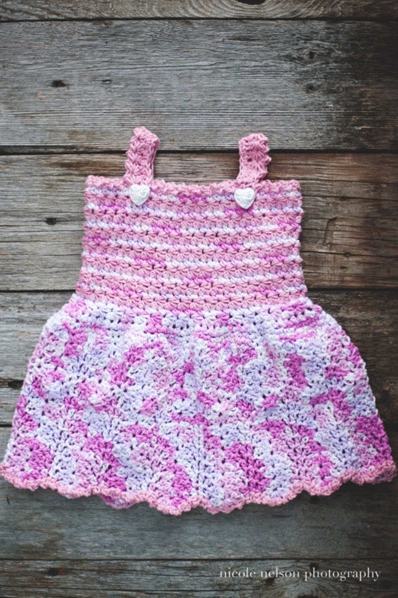 May include: A pink and white crocheted baby dress with a scalloped hem and two white heart-shaped buttons.