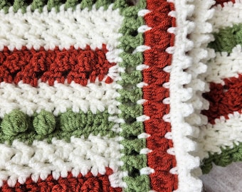 May include: A crocheted blanket with a white background and red and green stripes. The blanket has a scalloped edge and a textured pattern.