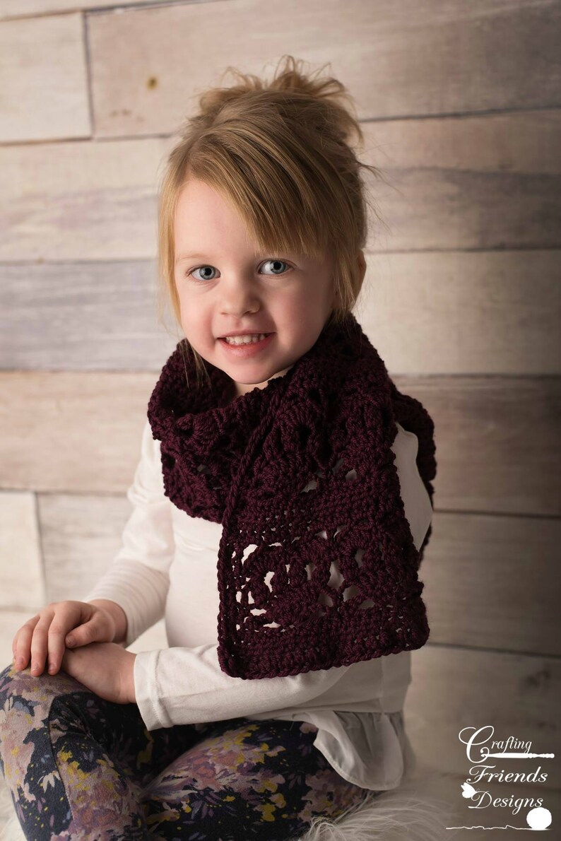 May include: A young girl wearing a white long-sleeved shirt and floral leggings is wearing a maroon crocheted scarf. The scarf has a lacy pattern and is tied around her neck.