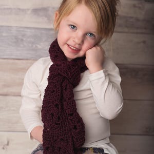 May include: A young girl with blonde hair wearing a white shirt and floral leggings. She is wearing a maroon crocheted scarf around her neck.