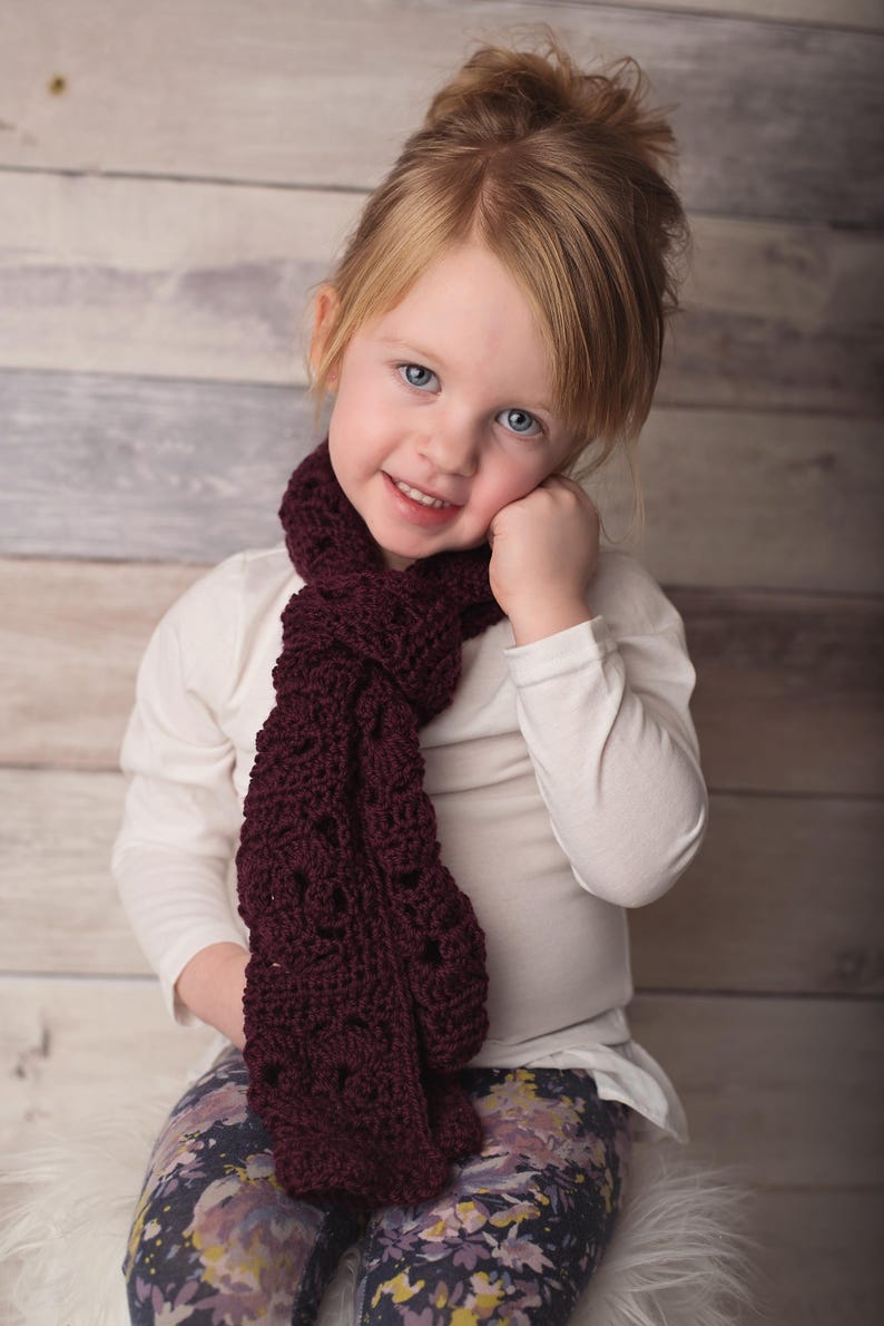 May include: A young girl with blonde hair wearing a white shirt and floral leggings. She is wearing a maroon crocheted scarf around her neck.
