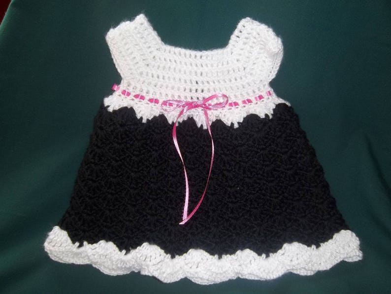 Peut inclure: Une robe de b&eacute;b&eacute; en crochet noir avec une bordure en crochet blanc et un ruban rose.