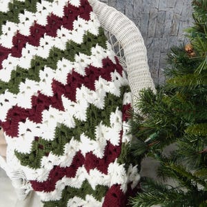 May include: A crocheted blanket with a chevron pattern in white, burgundy, and green. The blanket is draped over a white wicker chair and a small Christmas tree is in the background.
