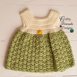 May include: A crocheted baby dress with a white top and green bottom. The dress has a yellow button in the center of the bodice. The dress is on a white wooden surface. Crafting Friends Designs.