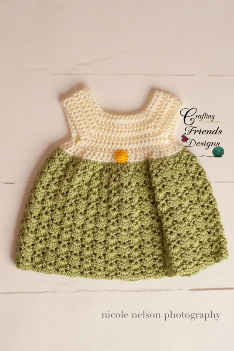 May include: A crocheted baby dress with a white top and green bottom. The dress has a yellow button in the center of the bodice. The dress is on a white wooden surface. Crafting Friends Designs.