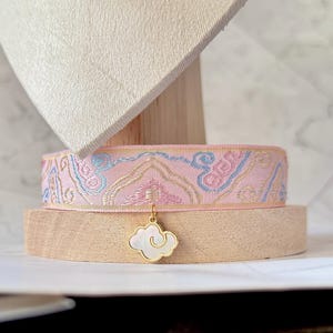 May include: A pink ribbon choker with a repeating pattern in blue, gold, and pink. A gold-colored cloud charm with a white center hangs from the choker. The choker is displayed on a wooden stand.