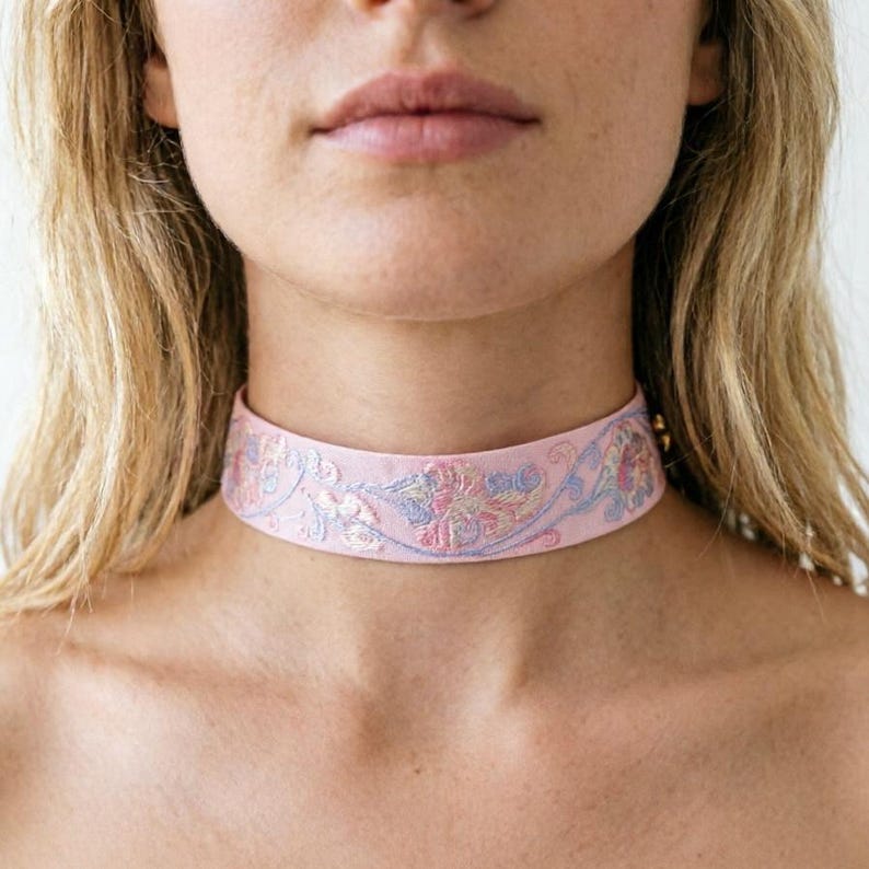 May include: A light pink choker necklace featuring an intricate embroidered floral pattern in shades of blue, yellow, and pink thread. The band is wide and sits flat against the neck, showcasing detailed stitching that creates a continuous botanical design.