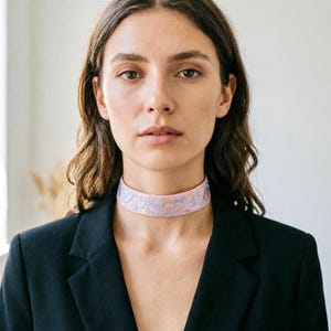 May include: A light pink choker necklace featuring a delicate, intricate floral pattern in shades of blue and white. The band is wide and sits flush against the neck, showcasing a detailed embroidery design that covers the entire surface of the fabric.