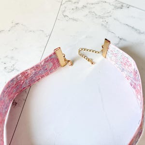 May include: A choker necklace featuring a wide, flat band with a textured, marbled pattern in shades of pink, blue, and white. The ends are finished with gold-toned metal clamps, and it includes a gold-toned chain extender with a small clasp.