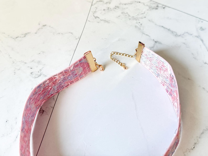 May include: A choker necklace featuring a wide, flat band with a textured, marbled pattern in shades of pink, blue, and white. The ends are finished with gold-toned metal clamps, and it includes a gold-toned chain extender with a small clasp.