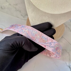 May include: A light pink fabric strap featuring an intricate woven floral pattern in shades of blue, gold, and magenta. The strap has a smooth texture and is held by a hand wearing a black glove, showcasing the detailed embroidery and design.