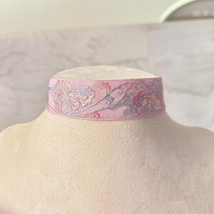 May include: A light pink choker necklace featuring an intricate embroidered floral pattern in shades of lavender, soft blue, and cream. The band is made of a woven fabric with a smooth texture, displaying a repeating botanical design across the front.