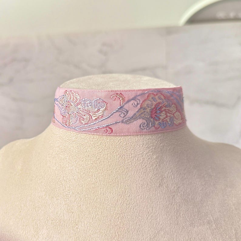 May include: A light pink choker necklace featuring an intricate embroidered floral pattern in shades of lavender, soft blue, and cream. The band is made of a woven fabric with a smooth texture, displaying a repeating botanical design across the front.