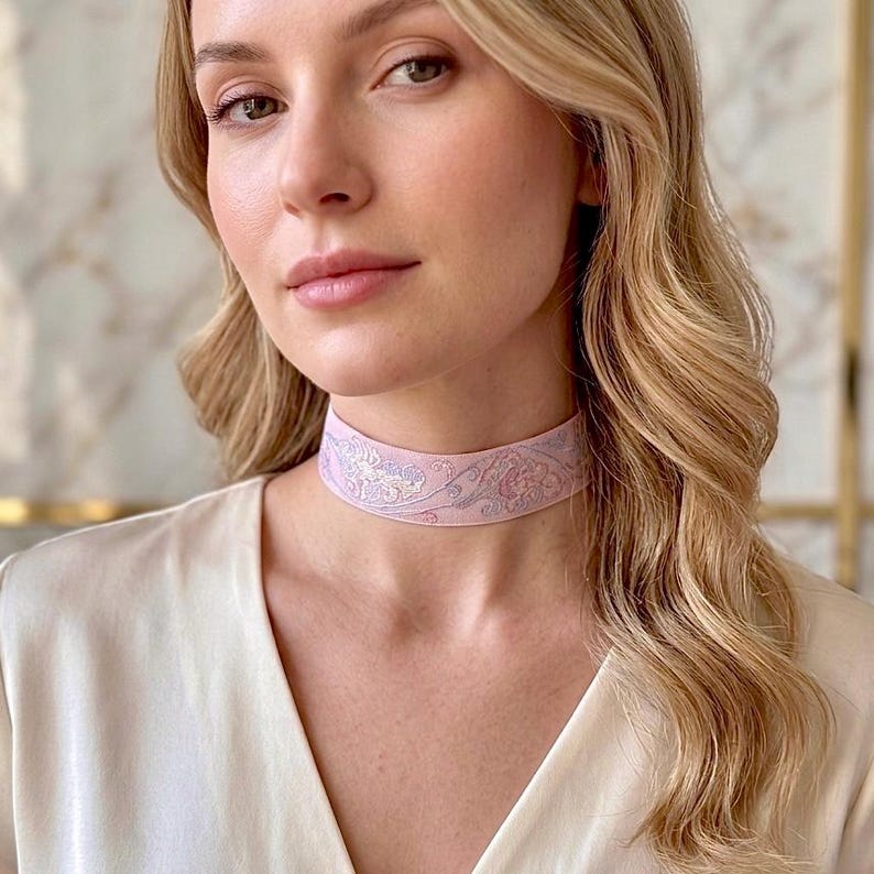 May include: A light pink choker necklace featuring a delicate, intricate floral embroidery pattern in shades of lavender and white. The band sits snugly around the neck, showcasing a detailed stitched design that spans the entire width of the fabric.