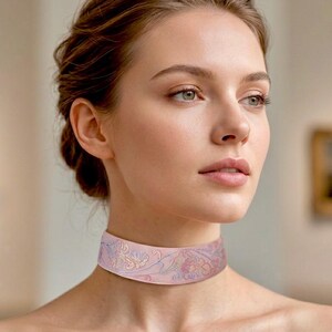 May include: A wide choker necklace featuring a soft pink fabric base with intricate, delicate embroidery in shades of light blue, lavender, and cream, forming a continuous floral pattern that wraps around the neck in a smooth, flat band.