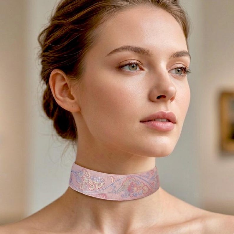 May include: A wide choker necklace featuring a soft pink fabric base with intricate, delicate embroidery in shades of light blue, lavender, and cream, forming a continuous floral pattern that wraps around the neck in a smooth, flat band.