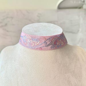 May include: A light pink choker necklace featuring a woven fabric band with an intricate floral pattern in shades of blue, purple, and cream. The design displays a repeating botanical motif across the surface of the textured textile material.