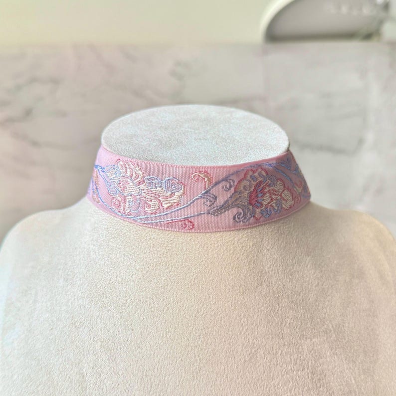 May include: A light pink choker necklace featuring a woven fabric band with an intricate floral pattern in shades of blue, purple, and cream. The design displays a repeating botanical motif across the surface of the textured textile material.