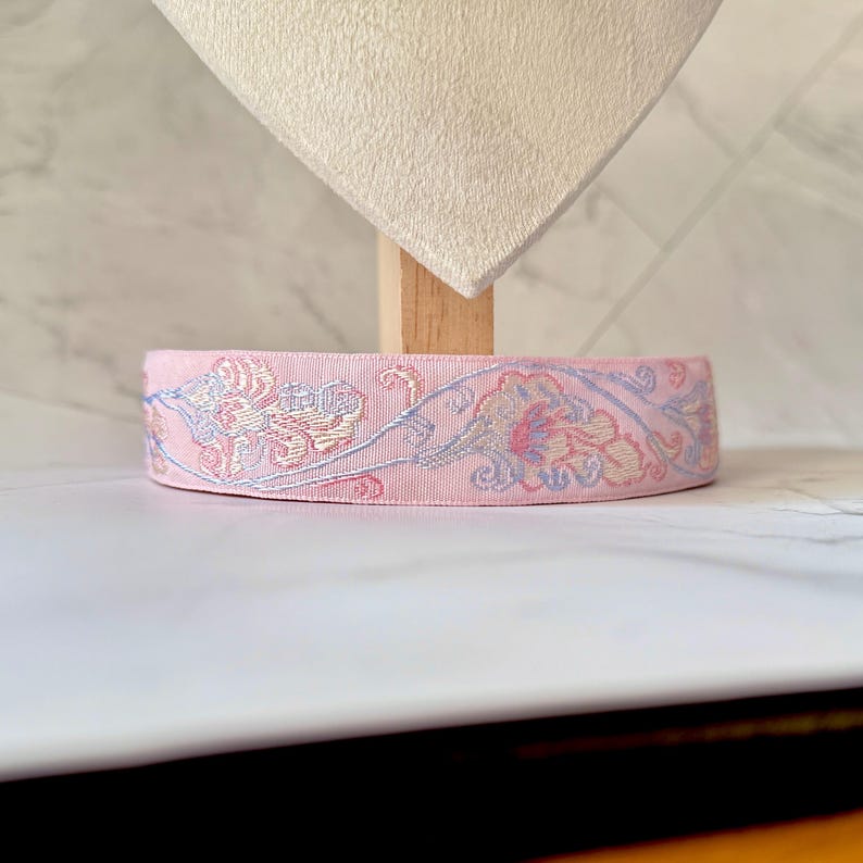 May include: A pink ribbon with a floral pattern in pastel colors. The ribbon is displayed on a white surface. The floral design includes shades of blue, pink, and yellow, creating a delicate and ornate appearance. The ribbon is likely used for crafting or decorative purposes.