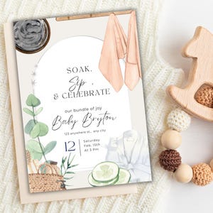 May include: A baby shower invitation with watercolor illustrations. The invitation features the text "SOAK, Sip, & CELEBRATE" and "Baby Bryton". The design includes images of spa elements, such as towels, cucumbers, and a robe. The date and time are also included.
