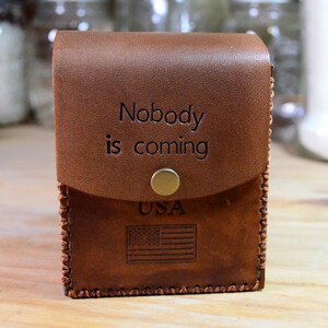 May include: A brown leather case with the words "Nobody is coming" embossed on the front. The case has a flap closure with a brass button and features the USA flag and text "USA" at the bottom.