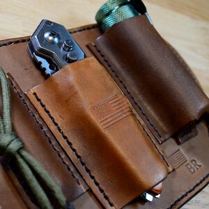 May include: A brown leather multi-tool organizer with three pockets. One pocket holds a silver folding knife, another a flashlight, and the third a pen with a green cord. The organizer has an embossed American flag.