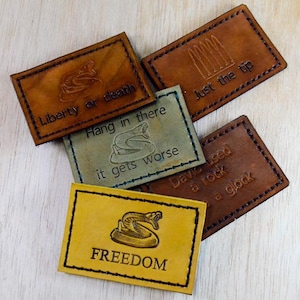 May include: A collection of rectangular leather patches in various shades of brown, green, and yellow. Each patch features a different engraved design and text, including a snake, bullets, and the words "Liberty or death" and "Freedom".