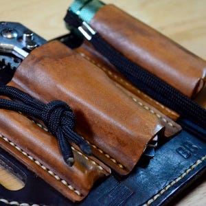 May include: A leather knife sheath with a black cord and a pocket knife. The sheath is brown and stitched with white thread. The knife is silver and has a black handle. The sheath is on a black leather base.