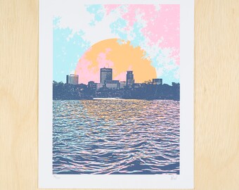 May include: A colorful print of a city skyline with a large orange sun setting over a body of water. The sky is a mix of pink, blue, and white.
