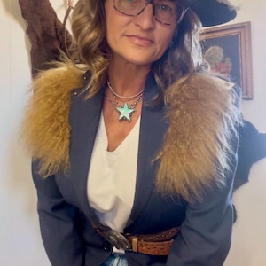 May include: A woman wearing a black hat, glasses, and a navy blazer with a fur collar. She is also wearing a white shirt, denim shorts, and a turquoise star pendant necklace. A brown belt with a decorative buckle completes the look.