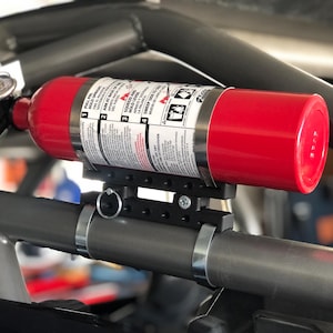 Könnte beinhalten: Ein roter Feuerlöscher, montiert an einem grauen Metallrahmen. Der Feuerlöscher wird mit schwarzen Halterungen und Metallklemmen befestigt. Anweisungen sind auf dem Etikett des Feuerlöschers sichtbar.