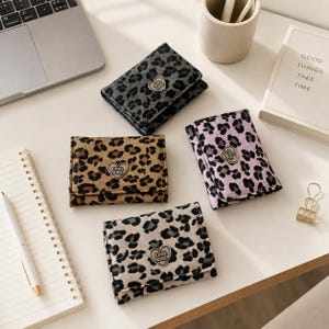 May include: Four rectangular wallets featuring leopard print patterns in gray, tan, pink, and beige. Each wallet has a central heart-shaped metal clasp engraved with the text COOL GIRL. The wallets are arranged on a flat surface alongside office supplies.