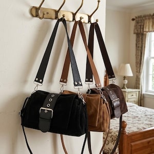 May include: Three shoulder bags hanging from a wall rack. Each bag features a suede-like texture, a wide adjustable strap, and a front flap secured by a large buckle with silver-toned metal eyelets. The bags are available in black, tan, and dark brown.