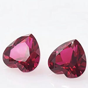 May include: Two loose heart-shaped gemstones with a deep magenta-red colour. Each stone features a faceted cut that creates multiple reflective surfaces, resulting in a brilliant appearance with sharp edges and a pointed base on a white background.