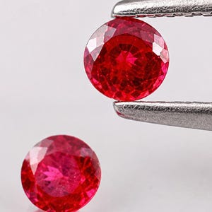 May include: Two round, faceted, vibrant red gemstones. One gemstone is held by silver-toned metal tweezers, while the other rests below it, displaying a clear reflection on the surface. The stones feature a brilliant cut with multiple facets.